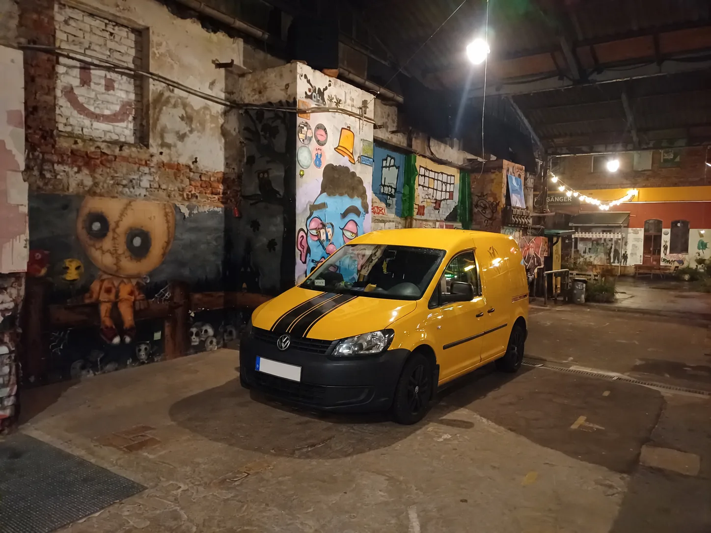 Ein gelber Van mit schwarzen Streifen auf der Motorhaube steht in einer alten Backsteinhalle mit Graffiti.